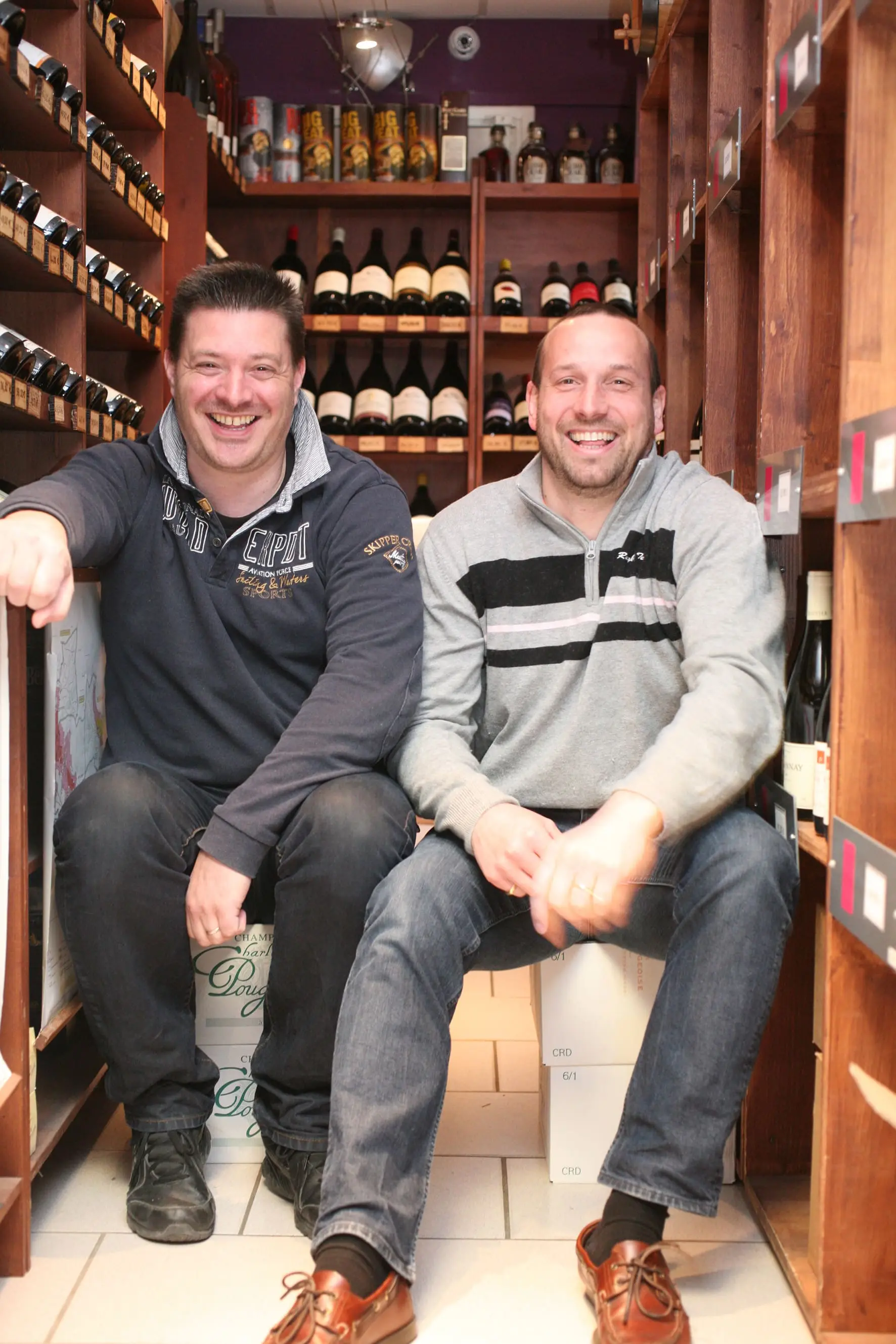 Deux patrons souriants dans leur boutique de vin, entourés de bouteilles.